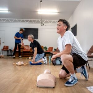 A CPR training session in progress, with three men practicing CPR techniques on manikins in a classroom setting. One man is kneeling and performing chest compressions, another is standing and assisting, and a third man is observing while kneeling