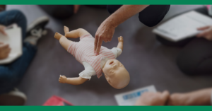 Close-up of a demonstration with an infant CPR manikin. A person points to the chest of the manikin while others take notes in the background.