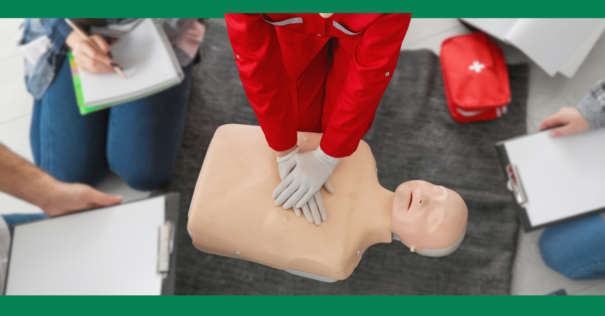 A first aid instructor is demonstrating chest compressions on a CPR mannequin while students take notes, emphasizing hands-on CPR training.